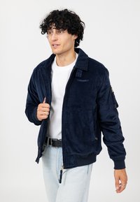 Marineblauwe corduroy bomberjack met een ritssluiting, ribbels aan de mouwen en een zijzak. Wit T-shirt eronder, lichtblauwe jeans.