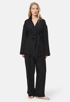 Vrouw staat blootsvoets, draagt een losse zwarte wikkeljas die ter hoogte van de taille is gestrikt en bijpassende wijde broek tegen een effen achtergrond.