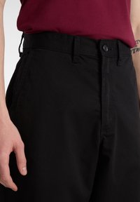 Pantaloni di cotone neri con passanti per cintura e chiusura con bottone, caratterizzati da una texture liscia e un design a gamba dritta. Una mano è appoggiata nei dintorni.