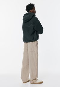 Fekete kapucnis dzseki, elasztikus alsó résszel, bézs kordbársony nadrággal párosítva. A modell oldalt áll, bemutatva a laza fazont és a textúrát.