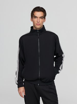 Giacchetto da tuta nero con zip, realizzato in tessuto liscio, caratterizzato da strisce bianche sulle maniche e un colletto alto.