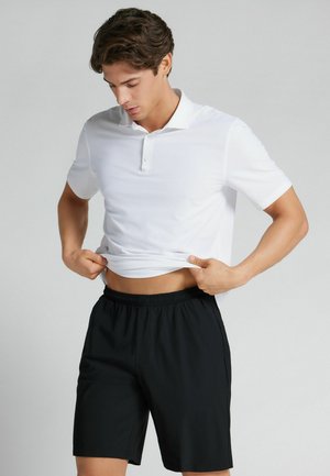 Ung mand iført hvid poloshirt og sorte shorts, løfter let sin skjorte og kigger ned, stående mod en ensartet lys baggrund.