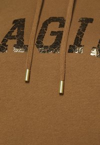 Gros plan d'un tissu marron avec des lettres noires texturées "AGI" et deux cordons marron avec des embouts métalliques dorés.