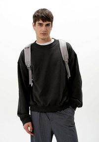Felpa nera con spalle cadenti e polsini a costine, indossata sopra una camicia bianca. Cinghie dello zaino grigio visibili sulle spalle.