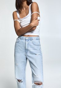 Donna che indossa un top bianco senza maniche con una spalla scoperta e jeans strappati azzurri, mentre tiene il braccio incrociato sul corpo.