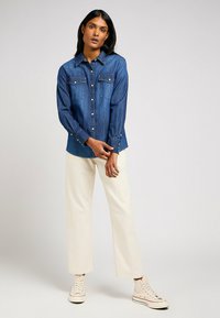 Camicia in denim blu scuro con colletto a punta, due tasche sul petto e bottoni argentati. Maniche lunghe con polsini a bottone, abbinata a pantaloni larghi color crema.