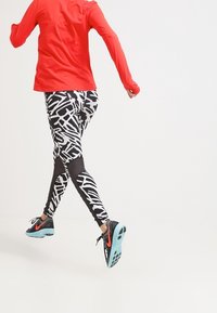 Maglia atletica rossa a maniche lunghe abbinata a leggings neri e bianchi con motivo e sneakers nere con suole turchesi, in una posa di corsa.