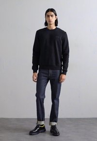 Sort sweatshirt med ribbede manchetter og kant, mørke jeans med opslåede manchetter og sorte sko med gule accenter. Neutral baggrund.