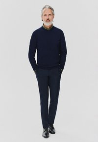 Granatowy sweter na zielonej koszuli z kołnierzykiem, w zestawieniu z ciemnymi spodniami i czarnymi butami. Model stoi z rękami w kieszeniach, na neutralnym tle.