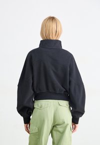 Schwarzer Fleece-Pullover mit hohem Kragen, überschnittenen Schultern und elastischem Saum. Kombiniert mit hellgrünen Cargo-Hosen mit Taschendetails.