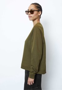 Mujer con cabello trenzado que lleva gafas de sol de carey, una sudadera verde oliva y pantalones negros, fotografiada de perfil contra un fondo liso.
