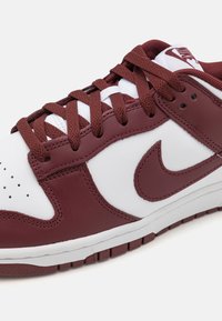 White and maroon leather sneaker with textured accents, perforated toe, flat maroon laces, and a white midsole, featuring a logo on the tongue.