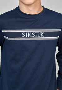 Camisa de manga larga azul marino, hecha de algodón, con un tejido texturizado y un logo SIKSILK blanco con dos rayas horizontales debajo.