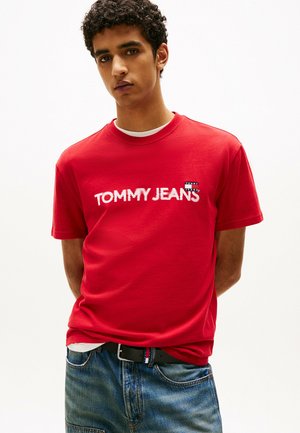 Rotes Baumwoll-T-Shirt mit dem Logo "TOMMY JEANS" in fettem weißen Text. Rundhalsausschnitt, kurze Ärmel, kombiniert mit blauen Denim-Jeans.