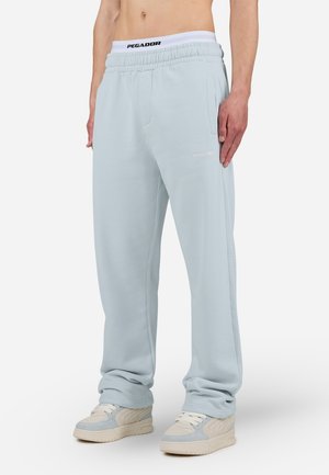 Man draagt lichtblauwe joggingbroek, witte tailleband met logo en beige sneakers, staand tegen een effen witte achtergrond.