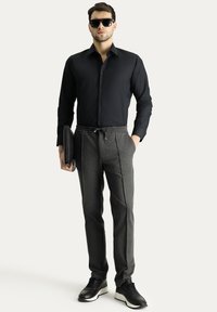 Camicia nera con bottoni indossata con pantaloni grigi con coulisse. I pantaloni in tessuto texture presentano tasche laterali. Sneakers nere unite completano il look.