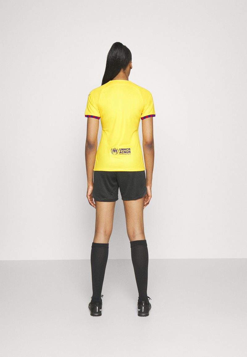 Camiseta deportiva amarilla de manga corta, pantalones cortos negros, calcetas negras hasta la rodilla y un logo del ACNUR en la parte posterior. Diseño simple, ajuste atlético.