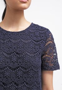 Femme portant un haut en dentelle bleu marine à manches courtes avec des motifs floraux et géométriques, décolleté et épaules visibles, sur fond blanc.