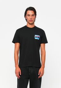Schwarzes Baumwoll-T-Shirt mit Rundhalsausschnitt und kurzen Ärmeln, das über einen mehrfarbigen grafischen Taschenaufdruck verfügt. Lässige Passform, keine sichtbaren Muster auf der Rückseite.