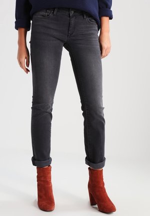 Persoon draagt donkergrijze slim-fit jeans met opgerolde boorden, gecombineerd met rode suède enkellaarzen, staand tegen een effen achtergrond.