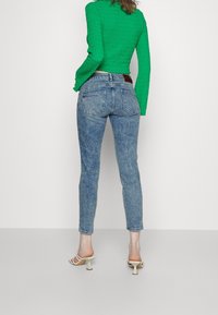 Vaqueros de mezclilla azul con cintura alta y ajuste slim, combinados con una blusa verde texturizada. Sandalias de tacón beige completan el look.