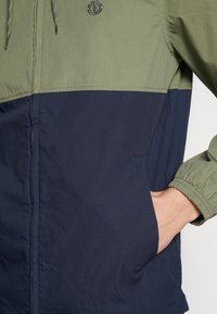 Grön och marinblå färgblockerad jacka med dragkedja framtill, elastiska muddar, justerbar huva med dragsko och sidfickor med en slät yta.