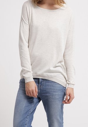 Maglione lungo beige chiaro a maniche lunghe con scollo rotondo. Presenta una texture morbida e polsini a costine, abbinato a jeans in denim blu.