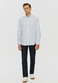 Jasnoniebieska koszula z długim rękawem, zapinana na guziki, z delikatnym wzorem, zestawiona z ciemnymi dżinsami i białymi trampkami, stojąca na jednolitym tle.