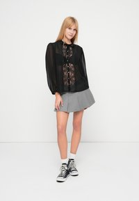 Zwarte doorschijnende blouse met kanten details, lange pofmouwen en knoopsluiting, gecombineerd met een grijze geplooide rok en zwarte sneakers.