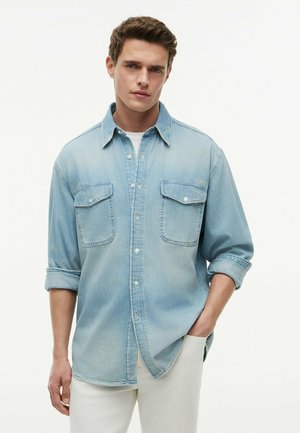 Ung mand iført en lyseblå denimskjorte med optrukne ærmer og hvide bukser, stående med den ene hånd i lommen mod en hvid baggrund.