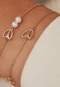 Goldkette mit Armband, das zwei runde Perlen und blattförmige Akzente zeigt, vor einem strukturierten Hautton-Hintergrund.