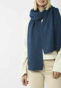 Dunkelblauder gestrickter Schal mit gerippter Textur. Leger getragen, umschlingt er den Hals und hat an der Unterkante ein Etikett.