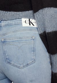 Jeans in denim azzurro chiaro con tasca posteriore a cucitura curva e etichetta bianca Calvin Klein Jeans, abbinati a un maglione lavorato a maglia nero e grigio.