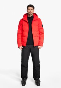 Rote Puffjacke mit Kapuze und gesteppter Optik, kombiniert mit schwarzen Hosen und Skistiefeln. Besitzt einen Reißverschluss und ein Logo-Patch am Ärmel.