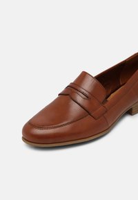 Mocassin pour femme en cuir marron avec petit talon empilé et détail de bride décorative sur le dessus, présenté sur un fond blanc.