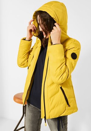 Femme ajustant la capuche d'une doudoune jaune vif avec une fermeture éclair noire et un écusson sur la manche, portant un pantalon foncé et un haut bleu marine.