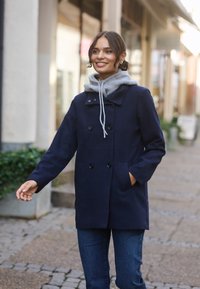 Marineblauer Wollmantel mit doppelt knöpfbarem Design, schwarzen Knöpfen und Seitentaschen, über einem grauen Kapuzenpullover mit Kordelzug getragen.