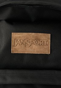 Μαύρο υφασμάτινο σακίδιο με ραμμένο ορθογώνιο καφέ σουέτ ταμπελάκι με την επιγραφή "JANSPORT" σε ανάγλυφα κεφαλαία γράμματα.