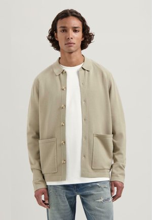 Beige knap-op jakke med krave, to forlommer og glat tekstur, båret over en hvid t-shirt og parret med slidte jeans.
