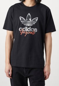 Černé bavlněné tričko s metalickým stříbrným logem Adidas a červeným textem "Originals" uprostřed, umístěné na jednobarevném pozadí.