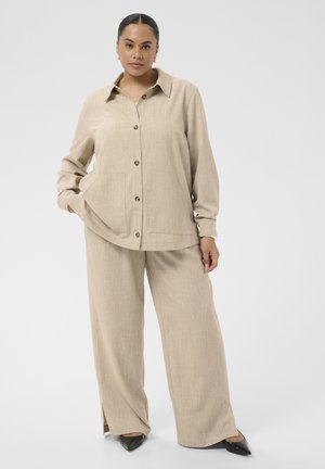 Femme portant une chemise beige boutonnée et un pantalon large assorti, avec des chaussures noires pointues, debout devant un fond uni.