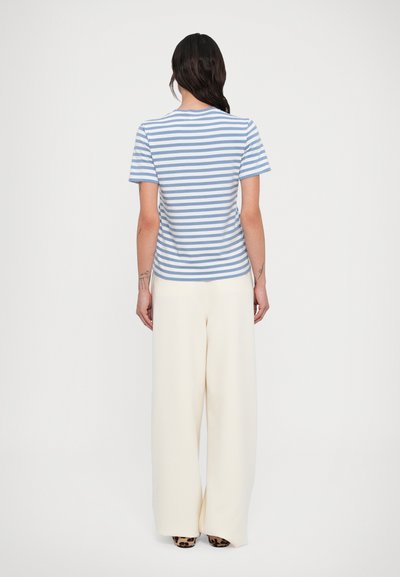 Γυναίκα που στέκεται με την πλάτη προς τον θεατή, ντυμένη με μπλε και άσπρη ριγέ κοντομάνικη μπλούζα, άσπρο παντελόνι με φαρδιά πόδια και παπούτσια με λεοπάρ μοτίβο.