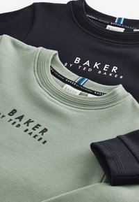 Na vrhu sta zloženi dva puloverja: en olivno zelen, drugi črn. Oba imata rebrast kroja in na sprednji strani natisnjen napis "BAKER by TED BAKER".