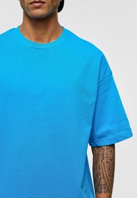 Helderblauwe katoen T-shirt met korte mouwen, ronde hals en een gladde textuur. Heeft een intrikat tatoeageontwerp op de onderarm.