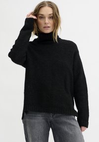 Schwarzer Rollkragenpullover mit strukturiertem Strick, lockerer Passform und langen Ärmeln, mit seitlichen Schlitzen am Saum. Kombiniert mit grauen Jeans.