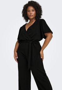 Juodas laisvalaikio kombinezonas su trumpomis rankovėmis, V formos iškirpte ir juosmens raišteliu. Minkšta medžiaga, tiesių kojų dizainas, šoninės kišenės.