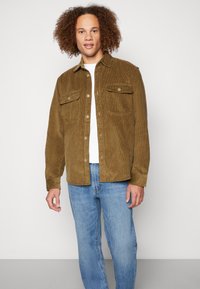 Brun kordfløyelsjakke med to brystlommer, knappelukking og en avslappet passform, båret over en hvit T-skjorte og blå jeans.