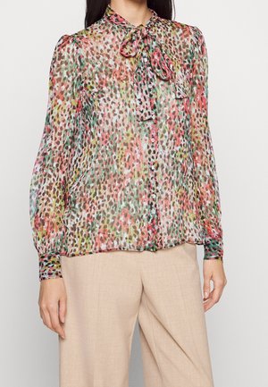 Frau trägt eine transparente, langärmelige Bluse mit mehrfarbigem Leopardenmuster und einer gebundenen Schleife am Hals, kombiniert mit beigen, weit geschnittenen Hosen.