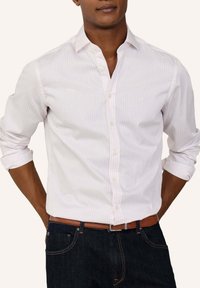 Mand iført hvid skjorte med lange ærmer med tynde røde striber, mørke jeans og brun læderbælte, hænderne bag ryggen.