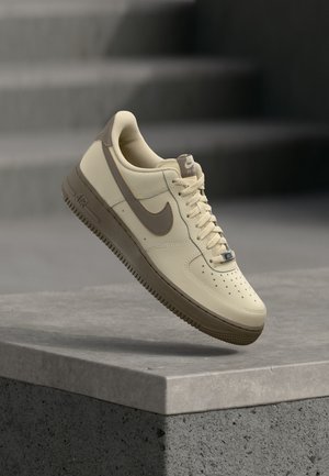 Bézs és barna Nike Air Force 1 sportcipő fűzőkkel, lebegve egy betonfelület felett, háttérben lépcsőkkel.